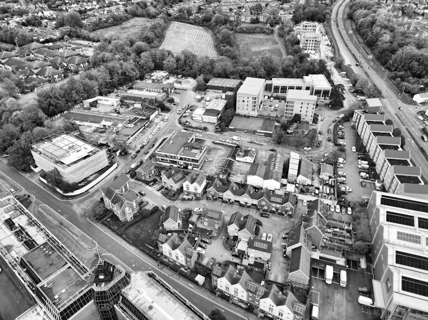 An aerial view in black and white showing a mixture of residential, commercial, and industrial buildings within a suburban area. In the foreground, there are rows of terraced houses with small driveways, some with parked cars visible. Adjacent to the houses, a narrow street separates the residences from a larger modern office or commercial building with flat roofs and a glass facade. Behind this building, a cluster of mid-rise office blocks or flats occupies part of the scene, with parking lots and vehicles around them. To the left, there is a larger, multi-storey industrial or warehouse structure with a flat roof and a surrounding paved area. Further back, a dense line of trees borders a field or park, and beyond that, a road curves into the distance alongside more greenery. The lighting appears natural with consistent illumination, and the overall scene depicts an urban or semi-urban environment suitable for services like private rubbish collection or waste management by companies such as Rubbish Collection Bromley.