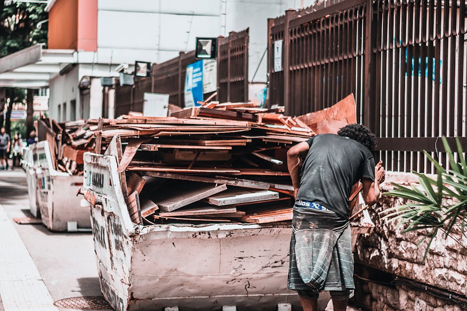 A man with curly dark hair wearing a dark T-shirt and checkered shorts is standing next to a large, overflowing skip filled with construction debris, including wooden planks, panels, and tiles. The debris appears rough-textured with a mix of brown, reddish, and gray tones, and some materials have a weathered or unfinished appearance. The skip, placed on a paved sidewalk, is situated close to a tall, dark brown metal fence with vertical bars, which runs along the edge of the property. In the background, there is a building with a white façade and orange accents, and some signage and streetlights are visible further down the pavement. The scene is illuminated by natural daylight, casting soft shadows, indicating an outdoor urban environment. This setting suggests a private or small-scale waste clearance activity, possibly related to home or garden renovation, aligning with the kind of on-site rubbish removal services provided by Rubbish Collection Bromley.