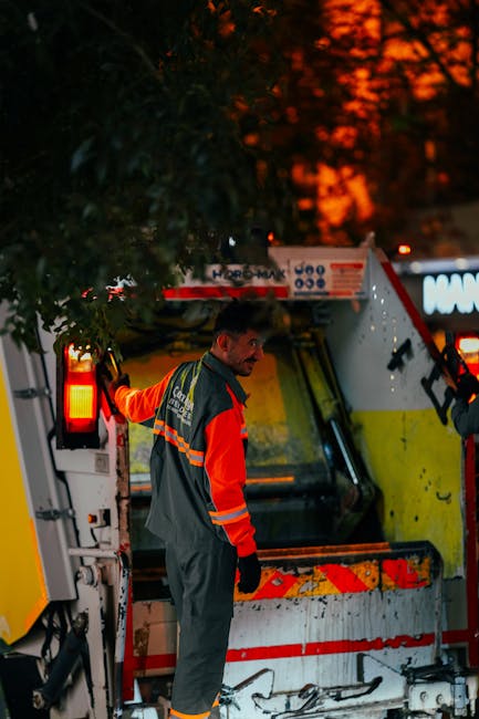 A waste management worker wearing a high-visibility orange and navy uniform stands outdoors beside a large industrial rubbish collection vehicle during dusk or early evening, with the sky illuminated by orange and red hues from the setting sun. The worker's face is visible, with short dark hair, and he is looking over his shoulder. The vehicle, primarily white with yellow and red reflective markings, has an open rear compartment revealing a cleaning or loading area, with some mechanical components and controls visible. The scene is set on a paved surface, possibly a street or driveway, with a background of trees and dark silhouette foliage. The ambient lighting highlights the reflective strips on the uniform and vehicle, emphasizing the practical aspects of private waste collection services, such as those offered by Rubbish Collection Bromley, in managing rubbish removal and garden clearance tasks in the Bromley area. The image captures the operational environment of independent rubbish disposal, emphasizing professional waste management practices in an urban setting.